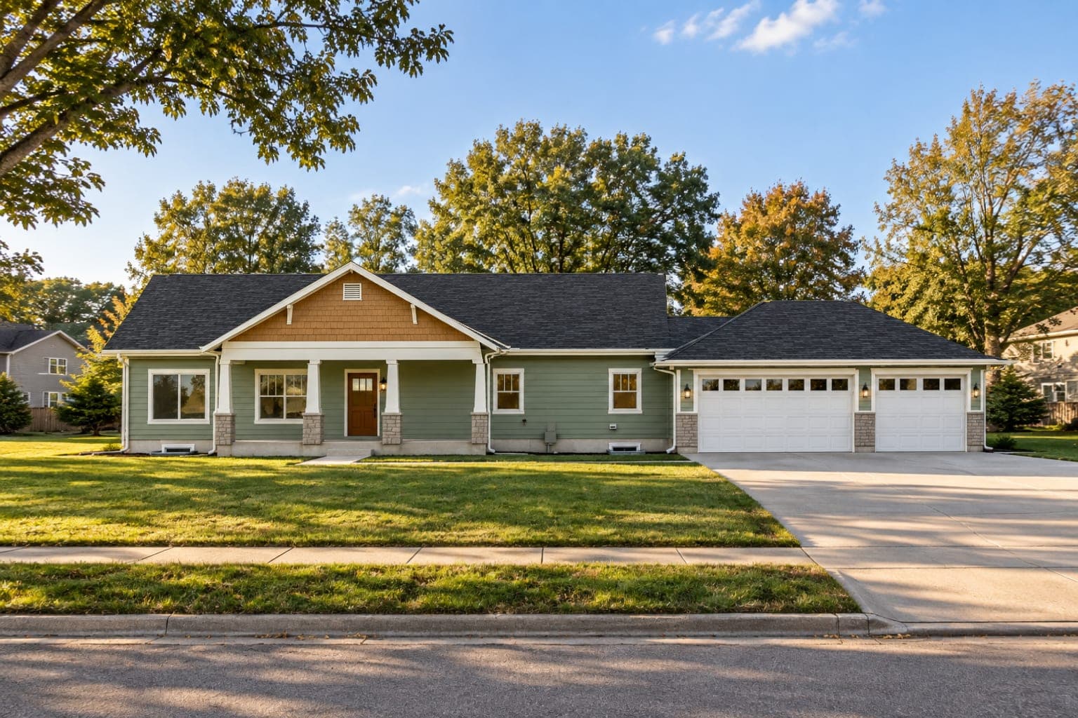 Front of a sage-green Craftsman ranch with a cedar shake gable, a covered porch on stone-based columns and a three-car garage