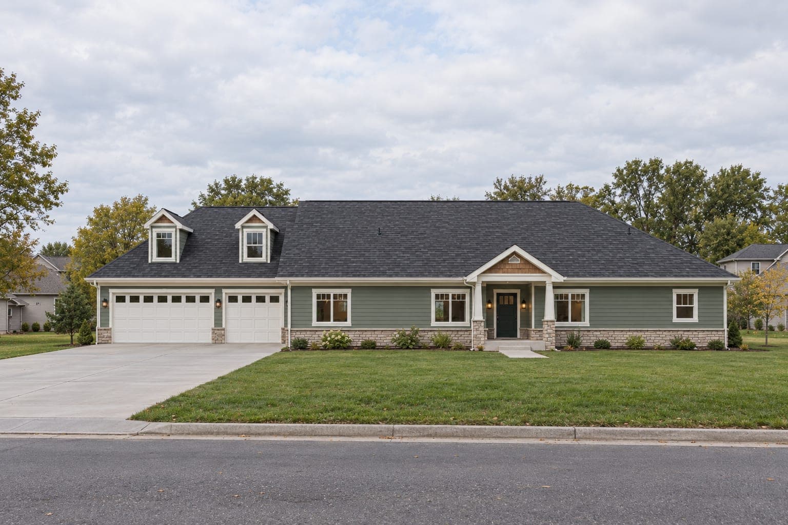 Front of a sage-green ranch with two roof dormers over a three-car garage, a stone wainscot and a gabled covered entry